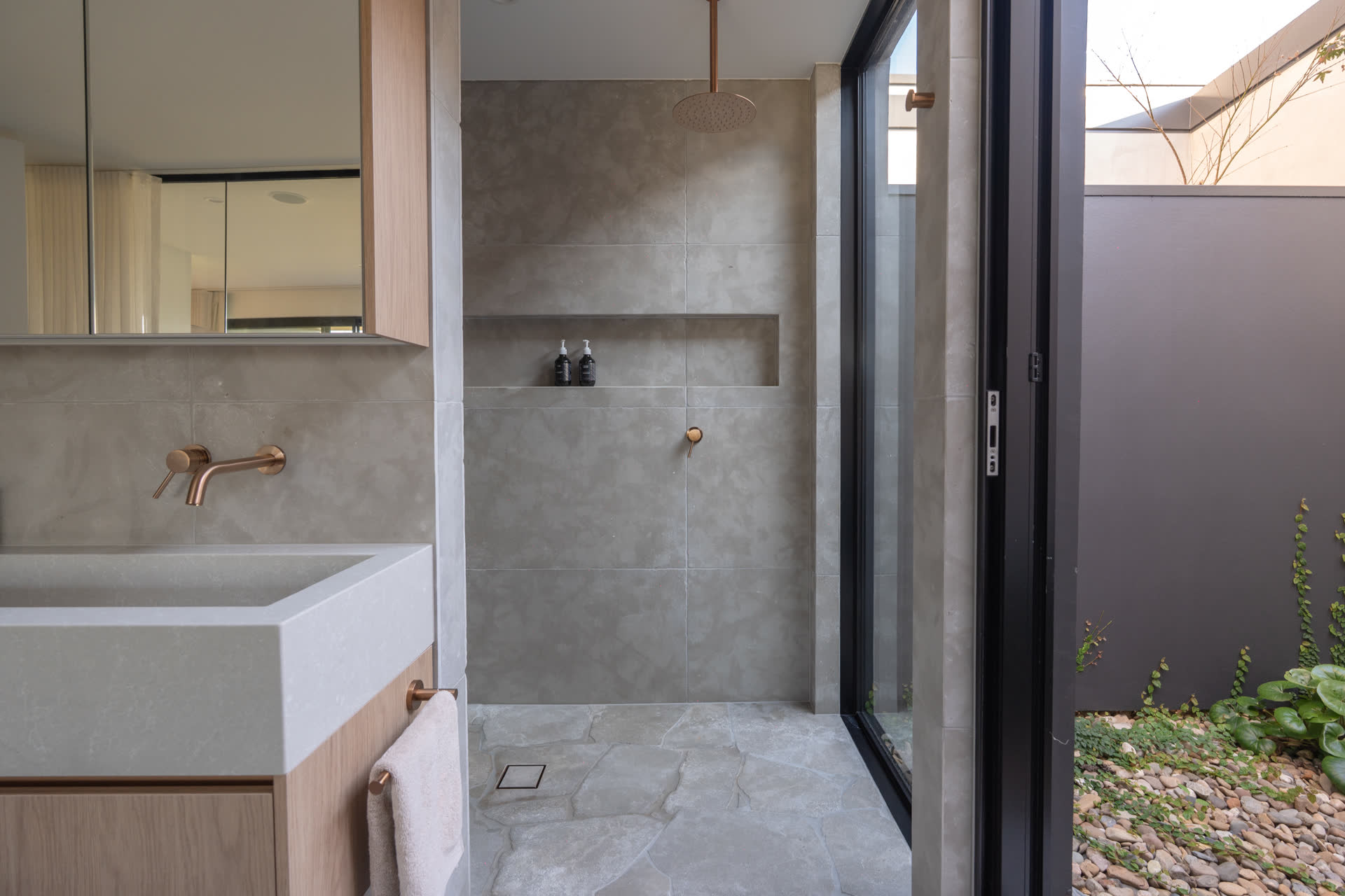Stone trough vanity and mirror cabinet inside the Glenorie ensuite, looking through full-height glazing to the outdoor stone tub in the courtyard