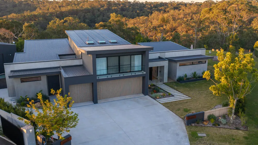 The front of the Glenorie house from above at golden hour — timber batten garage doors, entry path and the bushland ridge behind
