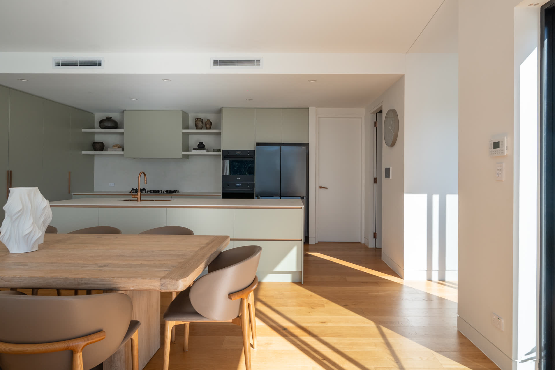 The Glenorie kitchen in afternoon light, seen across the oak dining table — island, brass tapware and sage joinery beyond