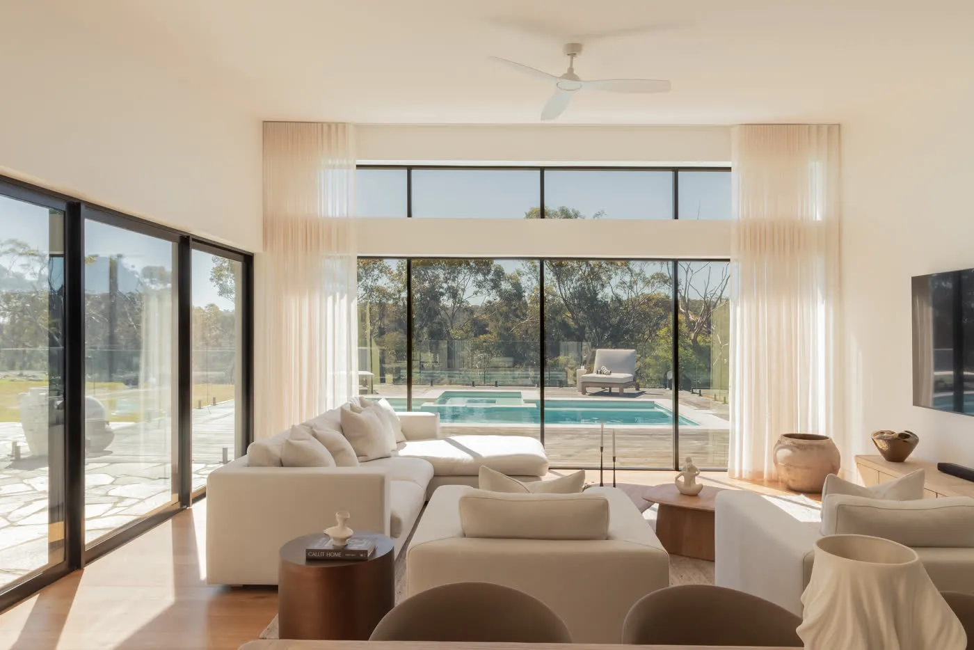 The Glenorie living room — white sofas and sheer curtains, full-height glazing holding the pool and bushland beyond